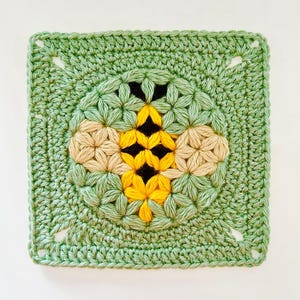 Puede incluir: Una pieza cuadrada de ganchillo hecha a mano con un diseño de abeja. La abeja está centrada en un fondo verde claro, con detalles amarillos, negros y beige. Los bordes del cuadrado tienen pequeños agujeros en cada esquina.