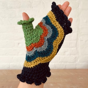 May include: A colorful crocheted fingerless glove with a green thumb cover. The glove has a black crocheted ruffle at the wrist and features a rainbow of colors including green, orange, red, blue, and yellow.