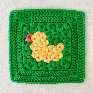 May include: A green crochet square with a yellow crocheted duck in the center. The duck has a red beak and a yellow body.