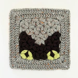 Puede incluir: Pieza cuadrada de ganchillo hecha a mano con un borde gris moteado. El centro presenta formas de flores grises, con acentos marrón oscuro y verde claro. La pieza muestra un patrón detallado y texturizado.