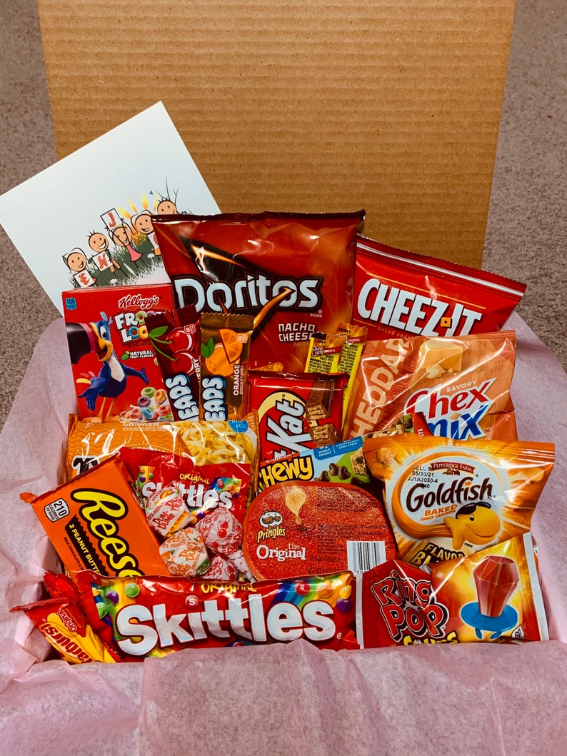 K&ouml;nnte beinhalten: Ein Geschenkkorb gef&uuml;llt mit einer Auswahl an Snacks, darunter Doritos, Cheez-Its, Skittles, Reese's und Goldfish Cracker. Der Korb enth&auml;lt auch eine Schachtel Frosted Flakes M&uuml;sli und eine Gru&szlig;karte. Die Snacks sind auf rosa Seidenpapier angeordnet.