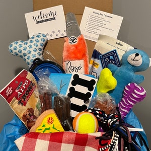 May include: A blue and white gift box filled with various dog toys and treats. The box includes a plush bone, a rope toy, a tennis ball, a chew toy, a dog treat, a water bowl, and a card that says "Welcome to the family".