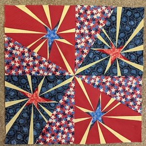 May include: A quilt block with a patriotic theme featuring a red, white, and blue starburst pattern. The block is made up of four different fabrics, each with a different design. The fabrics are sewn together in a way that creates a starburst effect.