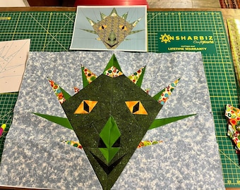 May include: A green and blue patchwork quilt block depicting a dragon face. The dragon has orange eyes, a black nose, and green spikes. The block is on a green cutting mat with a ruler and other fabric scraps.