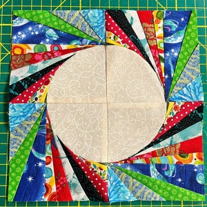 May include: A patchwork quilt block with a circular design in the center. The block is made up of various colorful fabrics, including red, blue, green, and black. The fabric is cut into strips and arranged in a spiral pattern around the center circle.