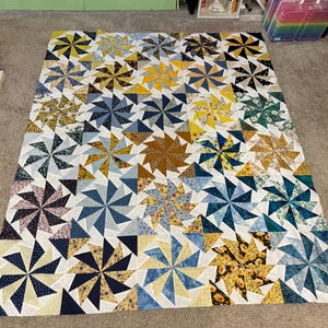 May include: A large, handmade quilt with a pinwheel design. The quilt features fabrics in shades of blue, yellow, brown, and floral patterns. The quilt is displayed on a neutral-coloured carpet.