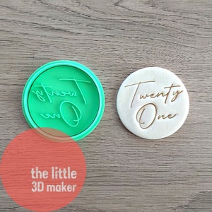 May include: A green plastic cookie cutter with the words "Twenty One" in a cursive font. The cutter is next to a white cookie with the same words imprinted on it.