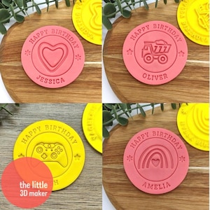 May include: Four round cookie cutters with pink and yellow handles. Each cutter has a different design: a heart, a truck, a video game controller, and a rainbow with a heart. The cutters are labelled with the names "Jessica", "Oliver", "Amelia", and "Happy Birthday".
