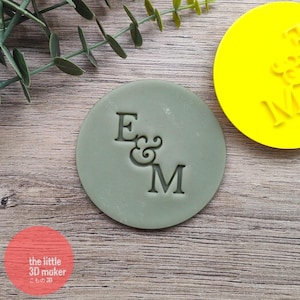 May include: A circular stamp in sage green, featuring the embossed letters "E & M". A bright yellow stamp is in the background. The image is set on a wooden surface, with a sprig of greenery in the upper left corner.