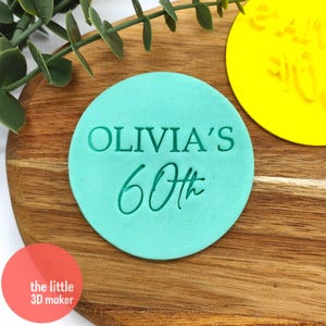 May include: A teal blue round cookie cutter with the text "Olivia's 60th" in white and green lettering.