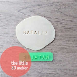 May include: A green plastic stamp with the word "JATAM" and the letters "LIE" on it. The stamp is used to imprint the name "NATALIE" into a piece of white dough.