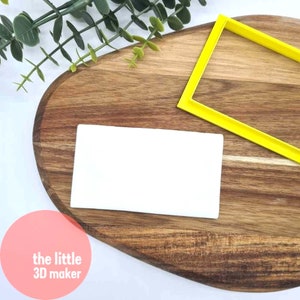 May include: A white rectangular silicone mould on a wooden surface. A yellow plastic frame is also on the surface. The text "the little 3D maker" is visible in the lower left corner of the image.