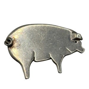 May include: A silver-toned metal brooch shaped like a pig in profile. The pig figure features a curled tail, a pointed ear, and a snout, with a smooth, slightly weathered surface texture and visible scratches across the body of the animal.
