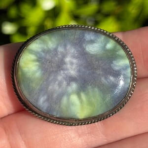 May include: Oval-shaped vintage brooch with a silver-tone rope-style border. The center features a marbled design in shades of blue, green, and yellow. The brooch is a classic accessory, suitable for adding a touch of vintage charm to any outfit.