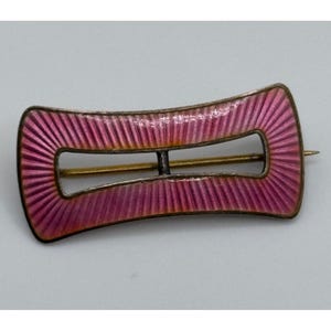 May include: A rectangular brooch with a central horizontal bar and a curved, concave frame. The surface features a vibrant pink enamel finish with a radiating, sunburst-like guilloché texture. A thin metal pin extends horizontally across the back.