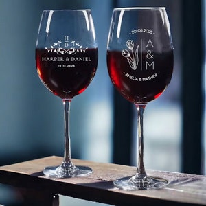 May include: Two clear wine glasses filled with red wine, personalized with names and dates. One glass has "Harper & Daniel" and "12.10.2026" etched on it. The other glass has "Amelia & Mathew" and "20.05.2025".