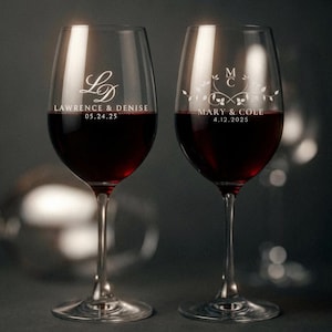 May include: Two clear wine glasses filled with red wine. Each glass is engraved with a monogram and names, along with a date. The glasses are set against a dark background, with soft lighting.