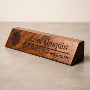 May include: A triangular wooden desk nameplate with a dark brown wood grain finish. The front face is engraved with a military rank insignia, the name Ariel Almquist, and the text 75FS Senior Enlisted Leader in a dark, recessed font.