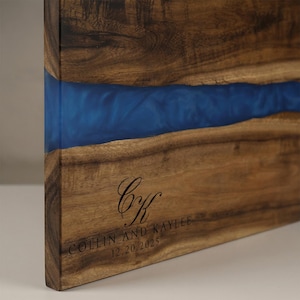 May include: Wooden serving board with a blue resin river. The board features a dark wood grain and is engraved with the initials "CK" and the names "COLLIN AND KAYLEE" along with the date "12.20.2025".