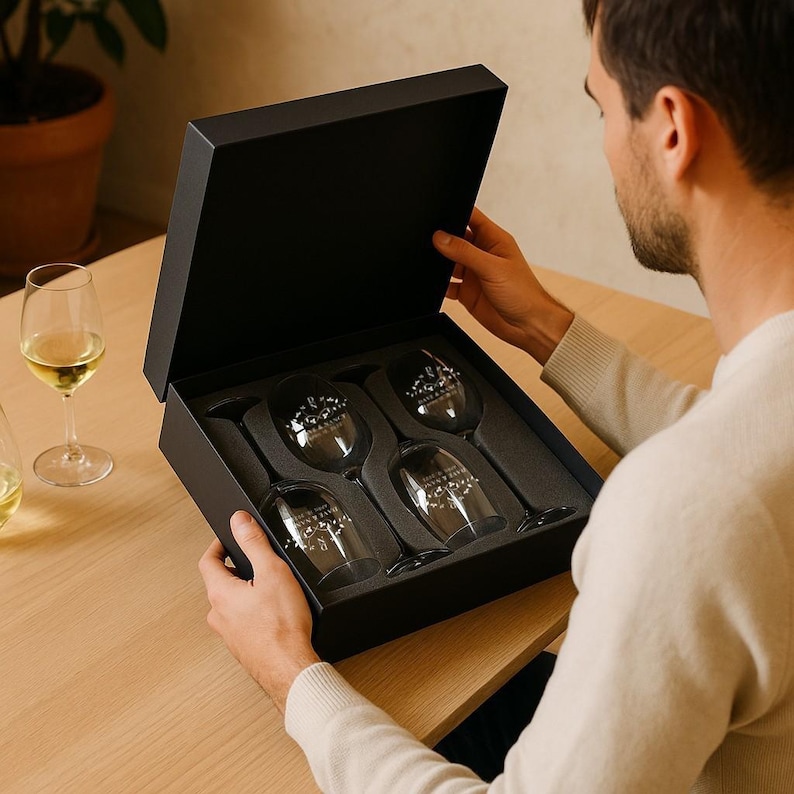 May include: A set of four clear wine glasses in a black gift box. Each glass has a decorative design. The box is open, revealing the glasses nestled in a foam insert. The glasses are on a wooden table.