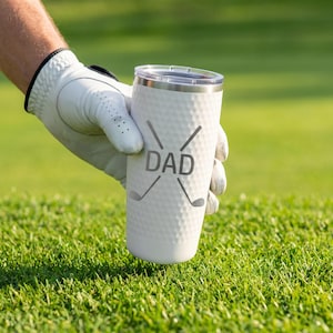 May include: White insulated tumbler with a dimpled texture, featuring the word "DAD" and golf club graphics. The tumbler has a silver rim and a clear lid. The background is a green golf course.