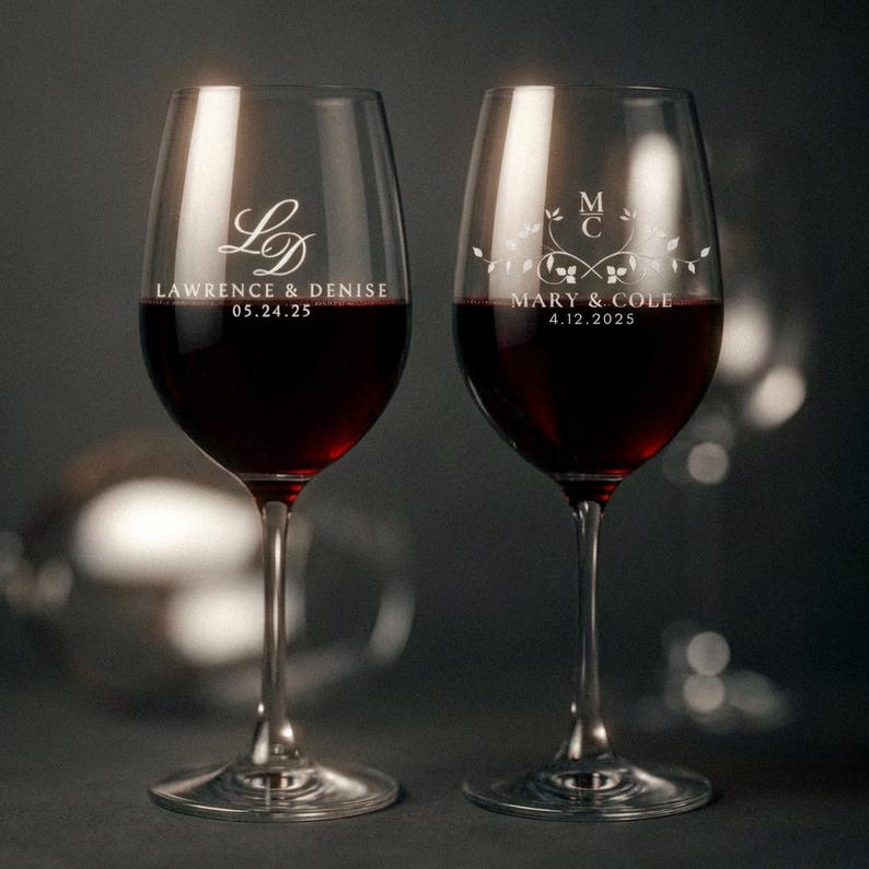 May include: Two clear wine glasses filled with red wine. Each glass is engraved with a monogram and names, along with a date. The glasses are set against a dark background, with soft lighting.