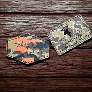 May include: Two camouflage patterned fabric patches. One patch is hexagonal and says "Alpha Wildlife Apparel". The other patch is rectangular and says "Est. 2024 Almonte Hunting Club".