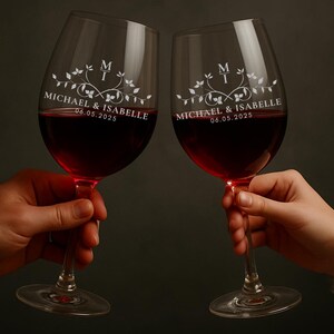 May include: Two clear wine glasses filled with red wine are held up in a toast. Each glass is engraved with a floral design, initials, and the names "Michael & Isabelle" along with the date "06.05.2025".