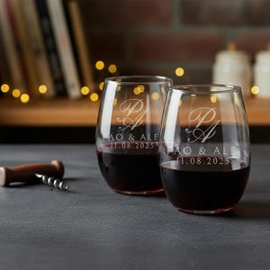 May include: Two stemless wine glasses filled with red wine. The glasses are engraved with the initials "P A" and the text "PAO & ALE 11.08.2025". A wooden corkscrew lies on the table next to the glasses. The background includes a shelf with books and string lights.