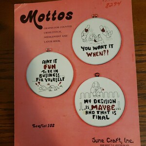 May include: A vintage leaflet titled "Mottos" for counted cross stitch, needlepoint, and latch hook. The cover is pink and features three circular designs with humorous sayings and illustrations. The leaflet is from June Craft, Inc.