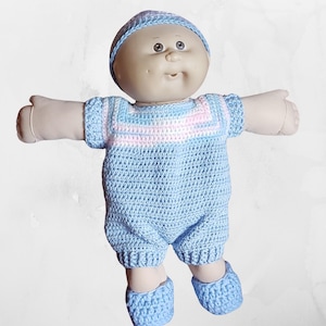 May include: A Cabbage Patch Kid doll wearing a blue crocheted outfit with pink stripes and a matching blue crocheted hat. The doll is wearing blue crocheted booties.