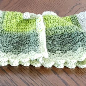 May include: A crocheted baby cardigan with a green, gray, and white color scheme. The cardigan has a scalloped edge and a button closure.