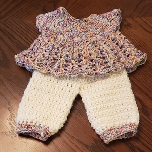 May include: A handmade crochet outfit for a baby, featuring a top and pants. The top is a mix of pastel colors, while the pants are a solid cream color. The outfit is made with a textured stitch, perfect for a baby.