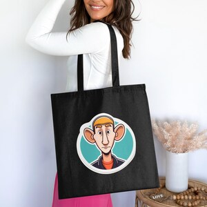 Peut inclure: Un sac fourre-tout noir orné d'un motif circulaire représentant un personnage de dessin animé aux grandes oreilles, au nez allongé et à l'expression neutre. Le personnage est sur fond bleu clair, portant une chemise orange et une veste sombre.