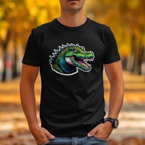 Peut inclure: Un t-shirt noir à manches courtes avec une impression graphique représentant une tête d'alligator verte avec un contour blanc. L'alligator a une apparence écailleuse texturée, des yeux jaunes et une gueule ouverte montrant ses dents.
