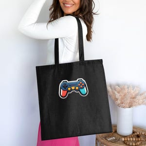 Peut inclure: Un sac fourre-tout noir orné d'une illustration graphique centrée représentant une manette de jeu vidéo. Le design de la manette comprend un corps bleu foncé, des boutons orange, ainsi que des poignées latérales bleu clair et rouge.