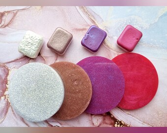 May include: Four small square containers of shimmery eye shadow in shades of white, gold, purple, and red, next to four round, larger containers of shimmery eye shadow in shades of white, copper, pink, and red.