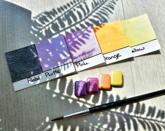 Könnte beinhalten: Ein Set mit fünf Aquarellfarben in den Farben Grau, Lila, Rosa, Orange und Gelb. Die Farben sind mit "Pastel", "Purple", "Pink", "Orange" und "Yellow" beschriftet. Ein Pinsel liegt auf einer Holzoberfläche.