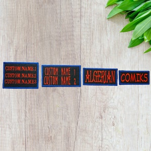 May include: Four black fabric patches with blue borders. The patches have white text that reads "CUSTOM NAME 1", "CUSTOM NAME 2", "CUSTOM NAME 1", "CUSTOM NAME 2". The fourth patch has red text that reads "ALGERIAN COMIKS".
