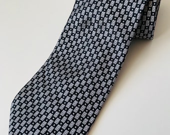 Puede incluir: Una corbata estampada en negro y plateado. La corbata presenta un diseño geométrico repetitivo de pequeños cuadrados y rectángulos. La corbata está doblada sobre sí misma, mostrando el patrón y la textura de la tela. Un accesorio clásico para vestimenta formal o de negocios.
