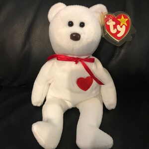 Puede incluir: Un peluche de oso Beanie Baby blanco con un parche de corazón rojo en el pecho y una cinta roja alrededor del cuello. El oso tiene una nariz marrón y una etiqueta en forma de corazón TY.
