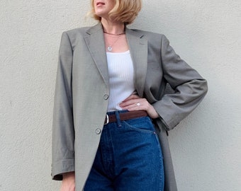 May include: A woman wearing a grey blazer over a white vest top and blue jeans. She is wearing a silver necklace and round glasses.