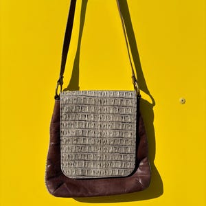 Könnte beinhalten: Eine braune Leder-Umhängetasche mit einer strukturierten, hellgrauen Klappe. Die Tasche hat einen langen, verstellbaren Riemen und ist vor einem leuchtend gelben Hintergrund platziert. Das Design der Tasche weist eine rechteckige Klappe und einen abgerundeten Boden auf.