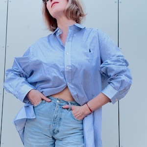 Peut inclure: Une chemise à rayures bleues et blanches avec un petit logo sur la poitrine, associée à un jean en denim délavé. La chemise est déboutonnée à la taille. La personne porte des lunettes de soleil et un bracelet rouge.