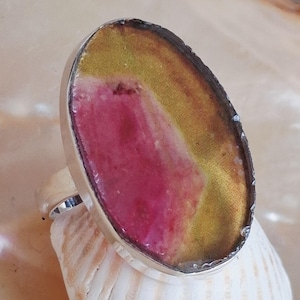 Könnte beinhalten: Ovaler Ring mit Silberband. Der Ring hat einen großen, farbenfrohen Stein mit Rosa-, Gelb- und Brauntönen. Der Stein ist in eine silberne Lünette gefasst. Der Ring liegt auf einer weißen Muschelschale.