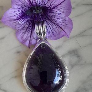 May include: A teardrop-shaped pendant with a deep purple gemstone set in a silver frame. The pendant is hanging from a silver chain.