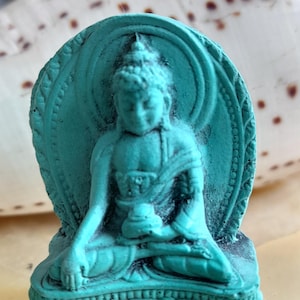 May include: A small turquoise blue statue of Buddha sitting in meditation pose. The statue is detailed with intricate carvings and is set on a base with a decorative design.