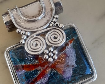 May include: Silver pendant with a square, multi-colored glass inlay. The glass has a sparkly, iridescent finish with shades of blue, orange, and purple. The pendant has a silver bail with a decorative design.