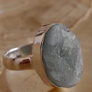 Könnte beinhalten: Ein Silberring mit einem großen, hellblauen, unregelmäßig geformten Edelstein. Der Stein hat ein strukturiertes, kristallines Aussehen und ist in einem polierten Silberring gefasst. Der Ring wird vor einem neutralen Hintergrund gezeigt.