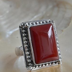 Könnte beinhalten: Ein silberner Ring mit einem rechteckigen roten Karneol-Edelstein, der in einem Perlenrand gefasst ist.